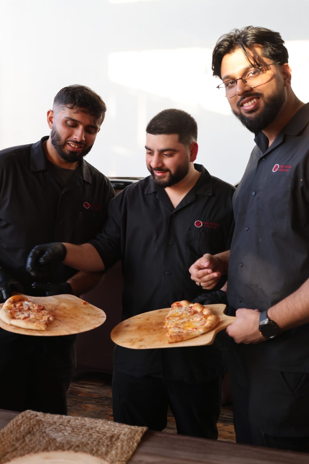 The three founders of The Pizza Station holding fresh pies on wooden peels — branded uniforms, Toronto